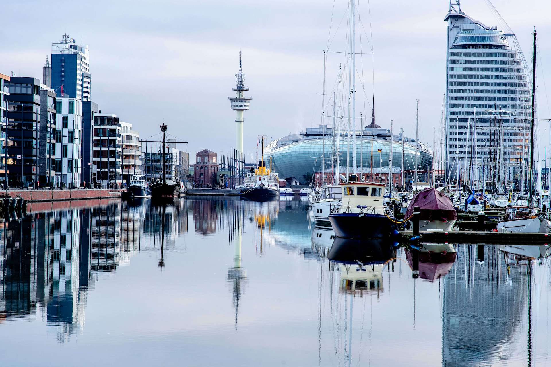 Blick auf den Hafen von Bremerhaven mit Segel- und Motorbooten, modernen Gebäuden, dem markanten Sail-City-Hotel und ruhigem Wasser mit Spiegelungen.