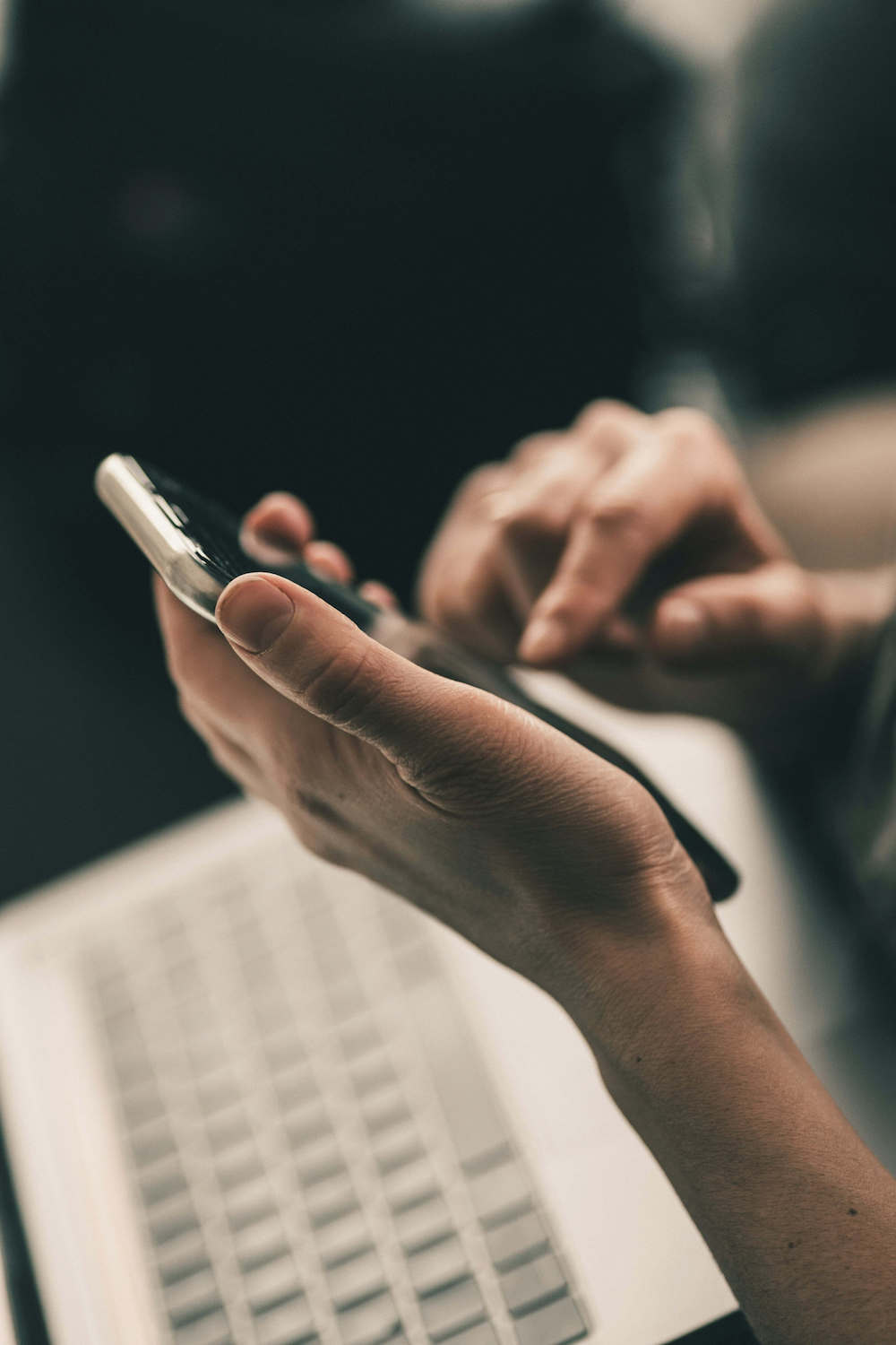 Hände bedienen ein Smartphone über einer Laptop-Tastatur.