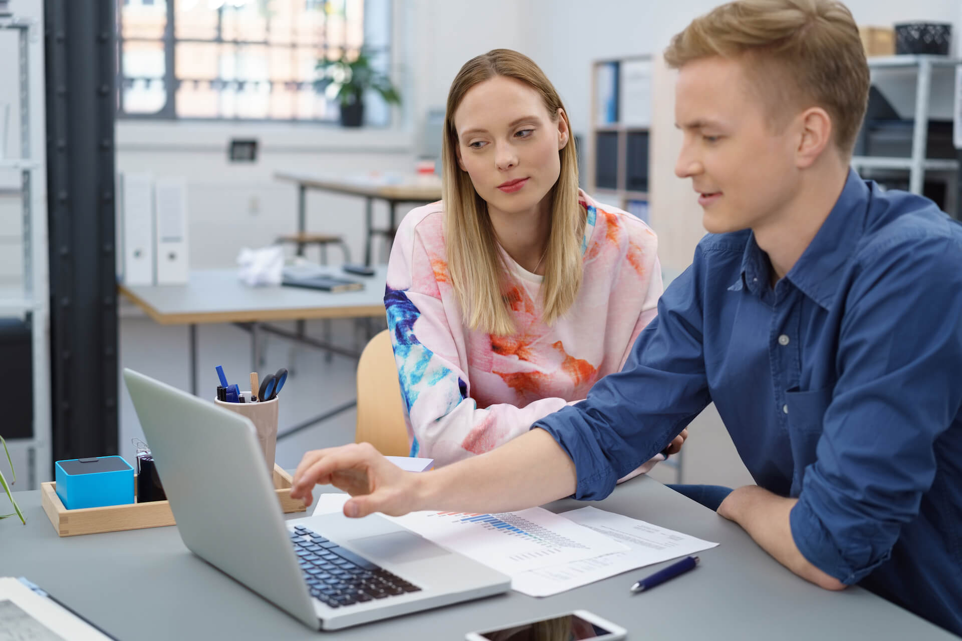 Zwei junge Menschen sitzen lächelnd an einem Schreibtisch mit Laptop und Unterlagen im Büro.